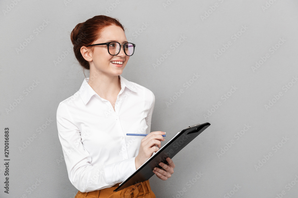 Portrait of pleased redhead businesswoman 20s wearing eyeglasses holding clipboard and smiling at camera in office