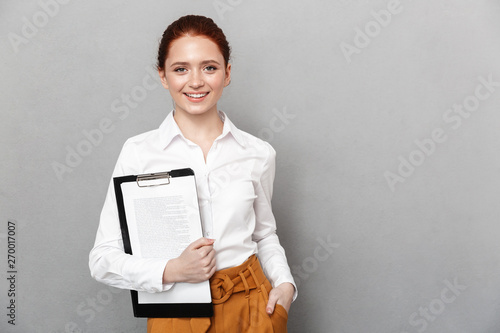 Portrait of cute redhead businesswoman 20s dressed in formal wear holding clipboard with documents and smiling at camera in office