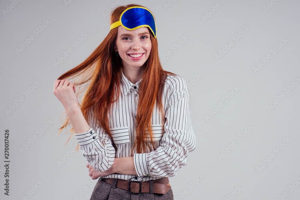 Good Morning. Smiling redhead young business woman in a striped shirt and blue yellow sleep mask white background studio Good Morning. Smiling redhead young business woman in a striped shirt and blue yellow sleep mask white background studio