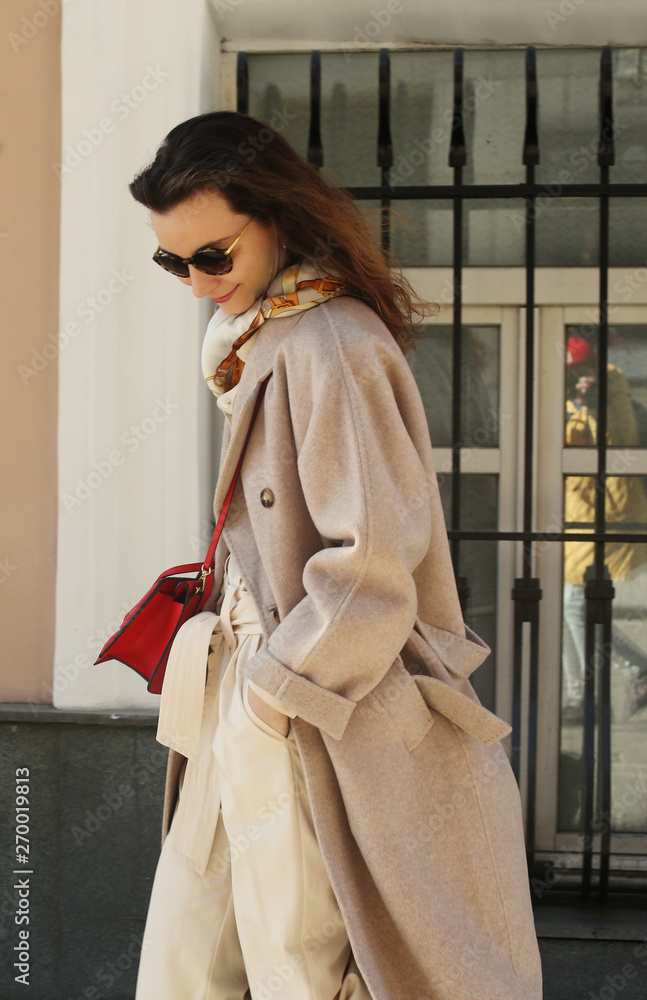 Portrait of a beautiful trendy fahsionista brunette with red lipstick in a beige coat with sunglasses. Posh stylish girl in Moscow. Black friday idea. Set sail champagne trendy color