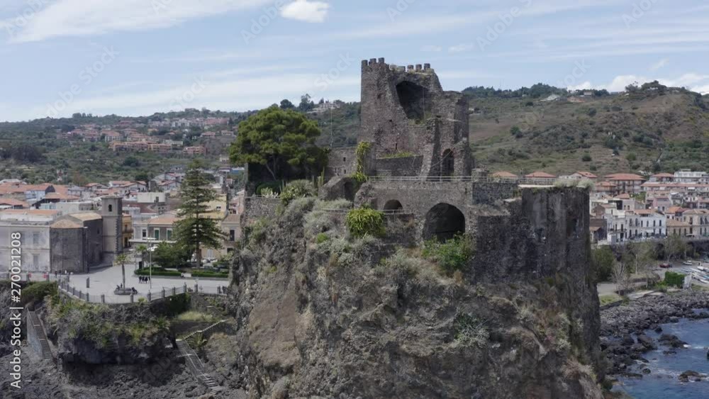 Drone shot of Aci Castello, Italy, with some trees, on the coastline with waves crashing on the shore, with the city and some hills behind it, on a cloudy day Drone shot of Aci Castello, Italy, with some trees, on the coastline with waves crashing on the shore, with the city and some hills behind it, on a cloudy day