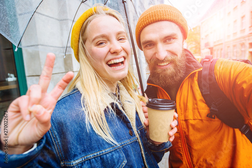 Self-portrait of nice cute lovely couple. Magnificent blond girl with wavy hair in casual denim jacket, yellow hat showing v-sign near eye hugging beard handsome man in orange hat and jacket outdoor. photo