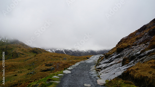 Stone path along an impactful view, surrounded by saturated grass, beautiful mountains and a foggy sky – captured during a hike at Snowdon in winter (Snowdonia National Park, Wales, United Kingdom)