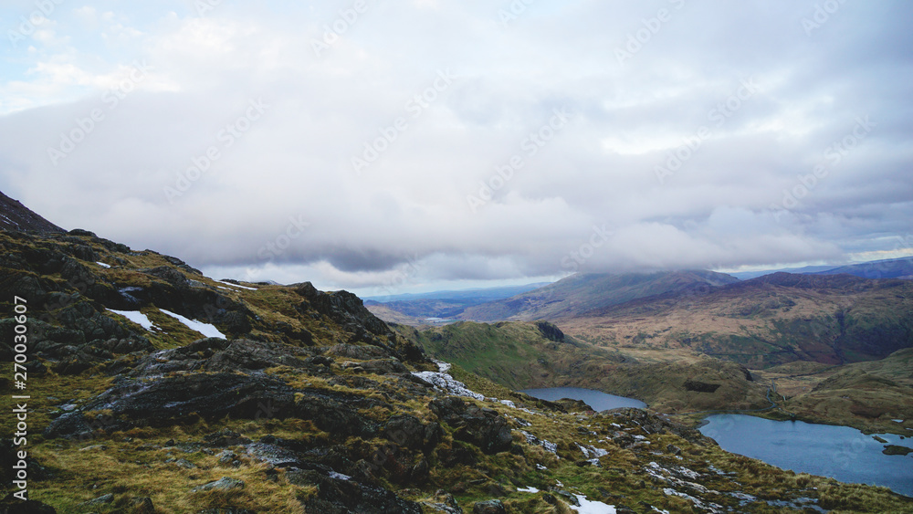 Stunning mountain view from the top seeing a lake, rugged valley and moody skies – captured during a hike at Snowdon in winter (Snowdonia National Park, Wales, United Kingdom)
