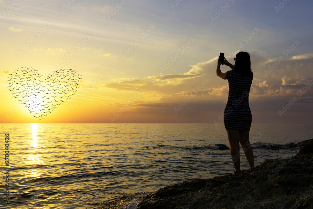 silhouette of a young girl. Photographs a beautiful sunset on the phone. heart of the silhouettes of birds in the sun.