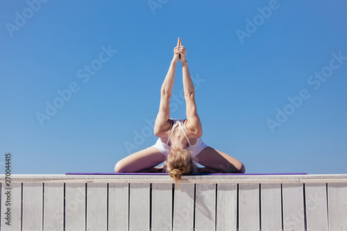 Sporty young woman sitting in variation of Upavishtha Konasana, Forward Bend posture. Slim girl practicing yoga outdoor, blue sky, white wooden terrace. Relax, healthy lifestyle concept photo