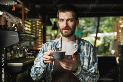 Portrait of brunette barista man holding cup of coffee while working in street cafe or coffeehouse outdoor