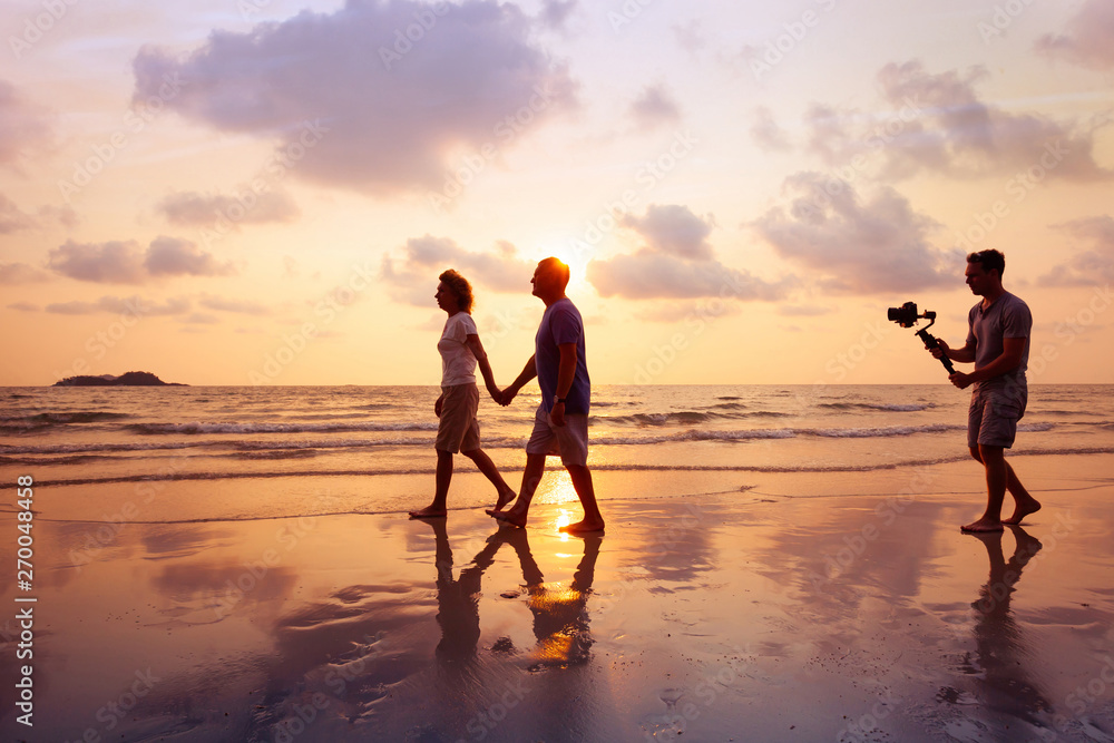 filmmaker filming video of couple with camera stabilizer on the beach at sunset, professional videographer working on film