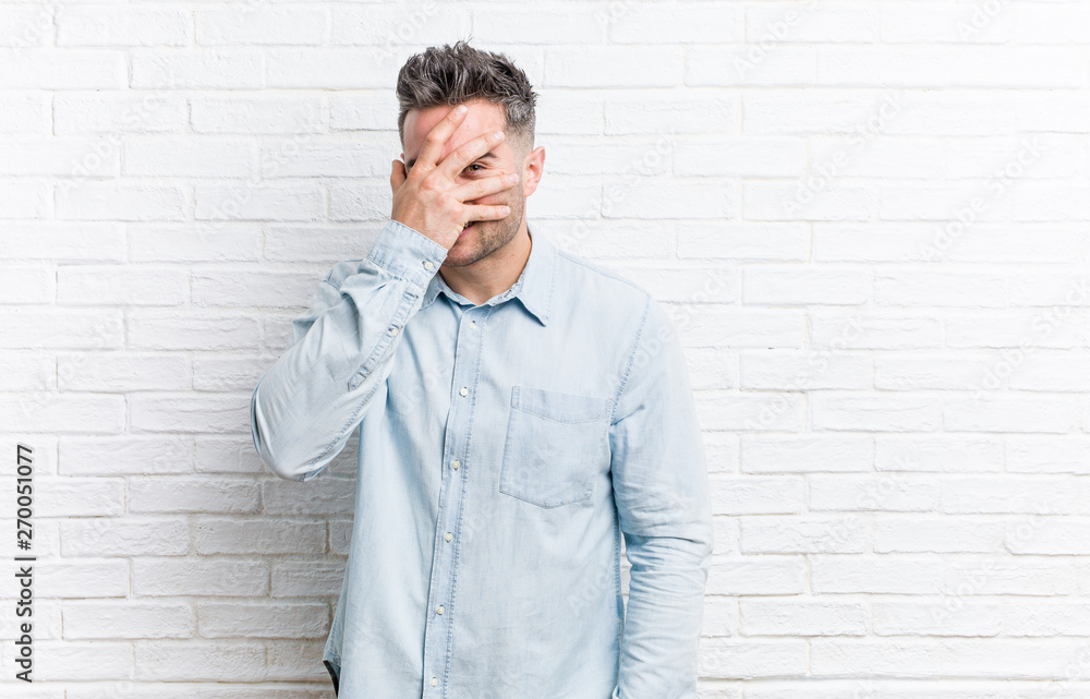 Young handsome man against a bricks wall blink at the camera through fingers, embarrassed covering face. Young handsome man against a bricks wall blink at the camera through fingers, embarrassed covering face.