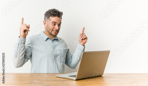 Young handsome man working with his laptop indicates with both fore fingers up showing a blank space.