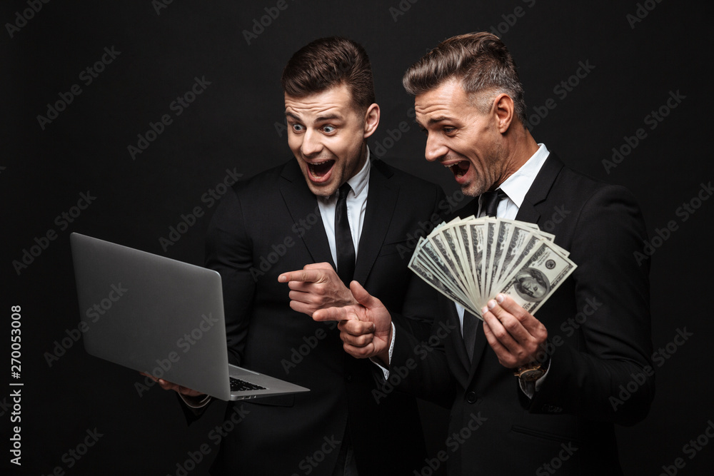 Portrait of excited caucasian two businessmen dressed in formal suit holding laptop and money banknotes Portrait of excited caucasian two businessmen dressed in formal suit holding laptop and money banknotes