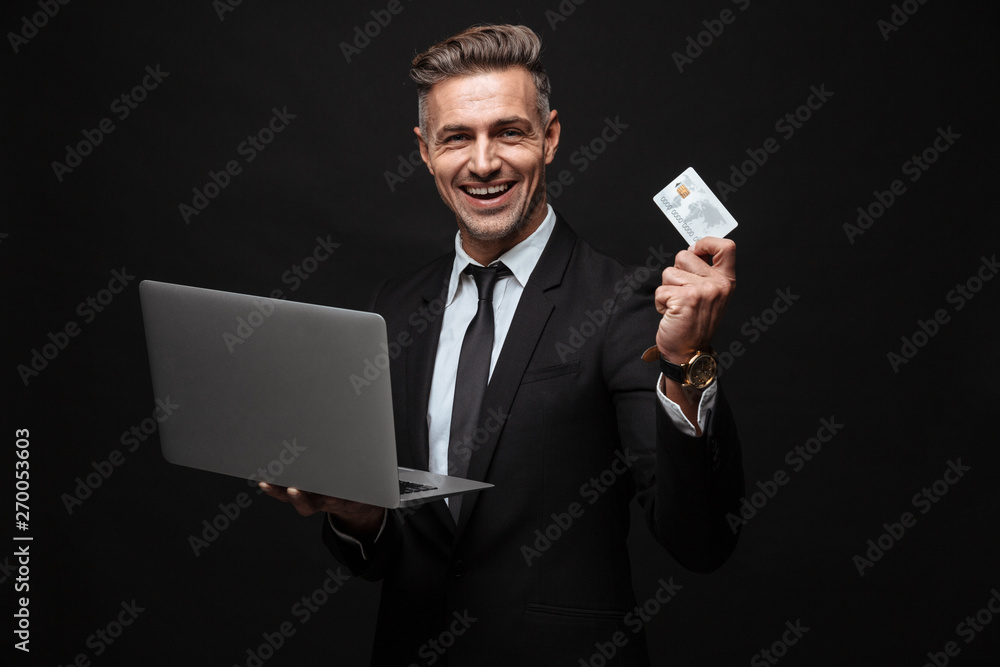 Portrait of manly smiling businessman dressed in formal suit holding laptop computer and credit card Portrait of manly smiling businessman dressed in formal suit holding laptop computer and credit card