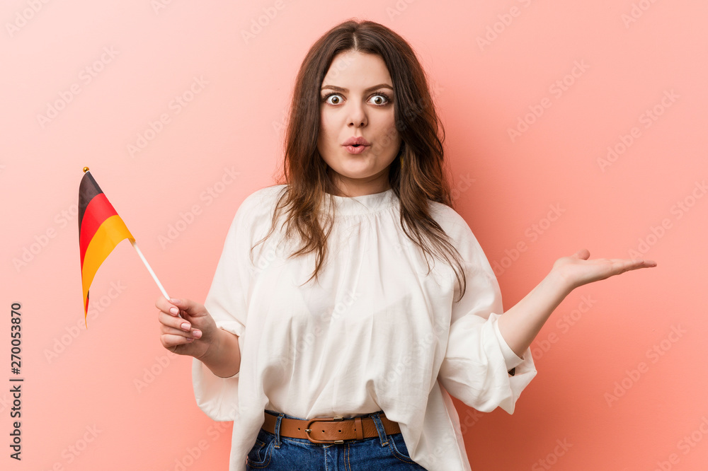 Young curvy plus size woman holding a germany flag impressed holding copy space on palm. Young curvy plus size woman holding a germany flag impressed holding copy space on palm.