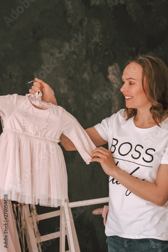 A beautiful smiling Woman in a white t-shirt and jeans is standing and holding a baby pink Princess dress on a dark background in the children's room