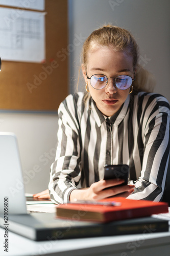 Photo of thinking caucasian woman architect wearing glasses using cellphone and sitting at workplace