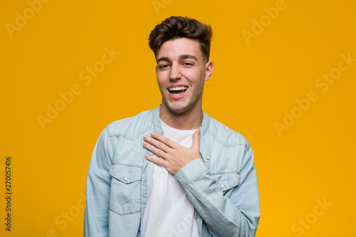 Young handsome student wearing a denim shirt laughs out loudly keeping hand on chest.