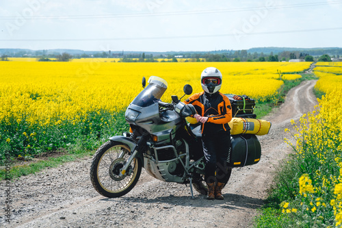 Girl in a protection outfit and glasses with touristic motorcycle. Rape yellow flowers field on background. Adventure trail tour, enduro and off road, summer day. extreme vacation concept