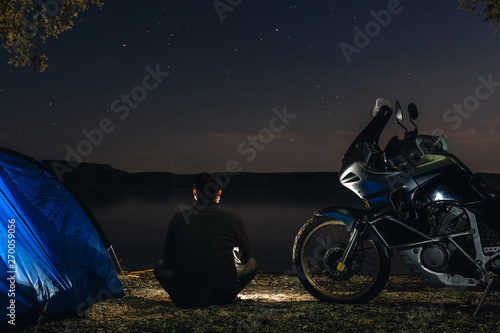 Man is sitting alone on beach in darkness and look othe sky Blue Camping Tent Illuminated Inside. Night Hours Campsite. Recreation and Outdoor. adventure motorcycle road trip
