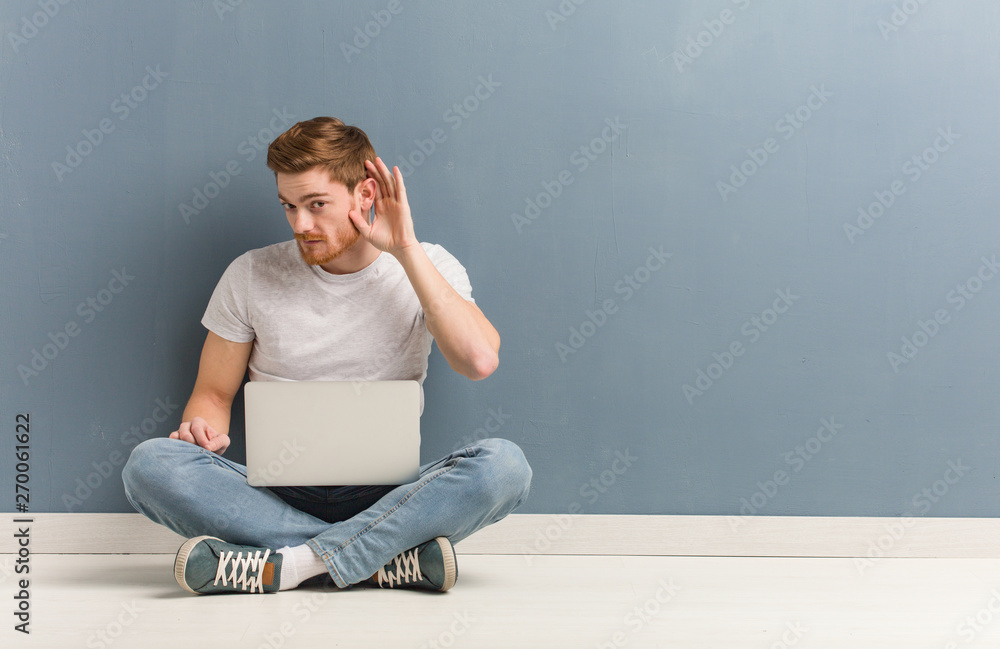 Young redhead student man sitting on the floor try to listening a gossip. He is holding a laptop. Young redhead student man sitting on the floor try to listening a gossip. He is holding a laptop.