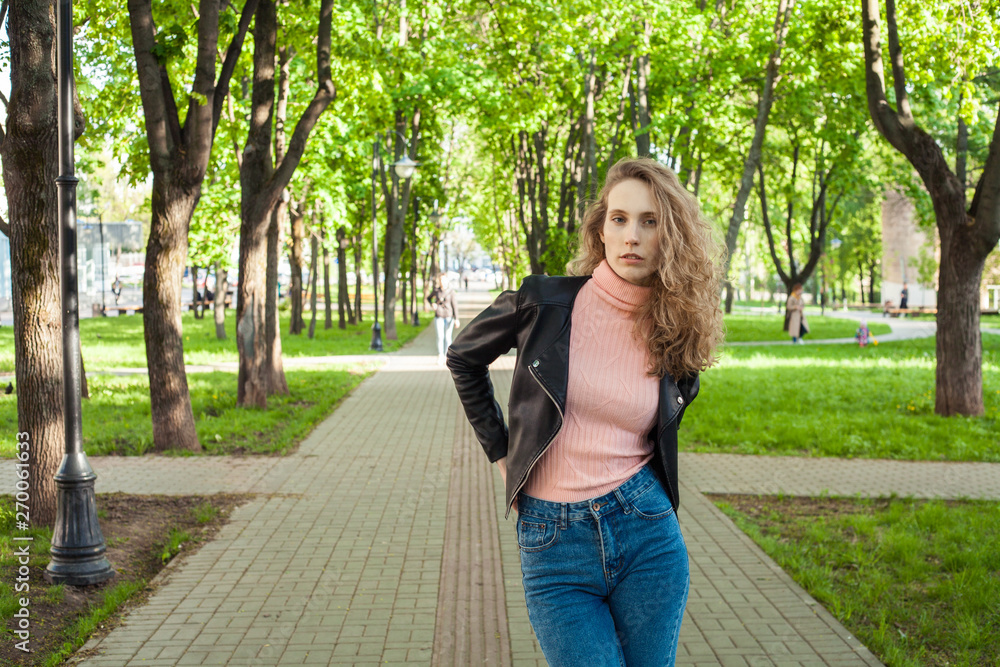 blonde girl with curly hair in blue jeans and a black leather jacket, standing in the Park on the background of the path and trees with a place for the inscription