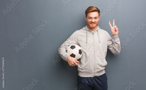 Young redhead fitness man fun and happy doing a gesture of victory. He is holding a soccer ball.