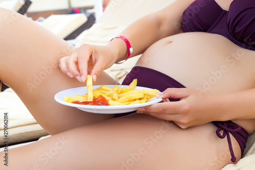 pregnant girl in a bathing suit eating fast food fried potatoes on a sunbed by the pool. Concept unhealthy food and vacation holiday