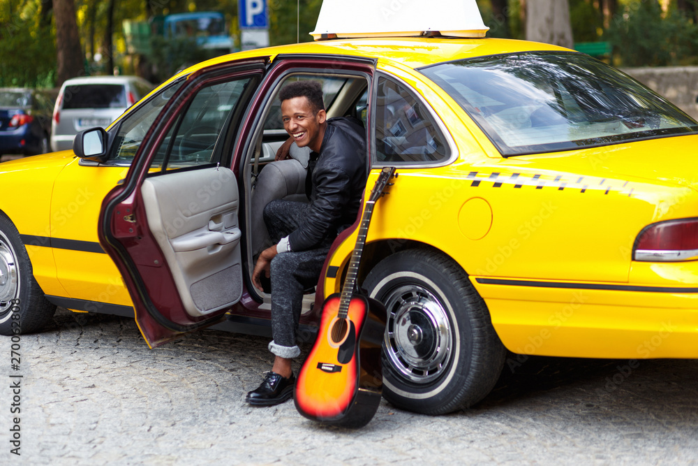 Happiness guy seated in interiors of taxi, looking a side and smiling,with left leg outside, near guitar, in street.