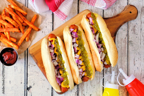 Hot dogs topped with onions, relish, mustard and ketchup served with french fries. Top view table scene on a white wood background.