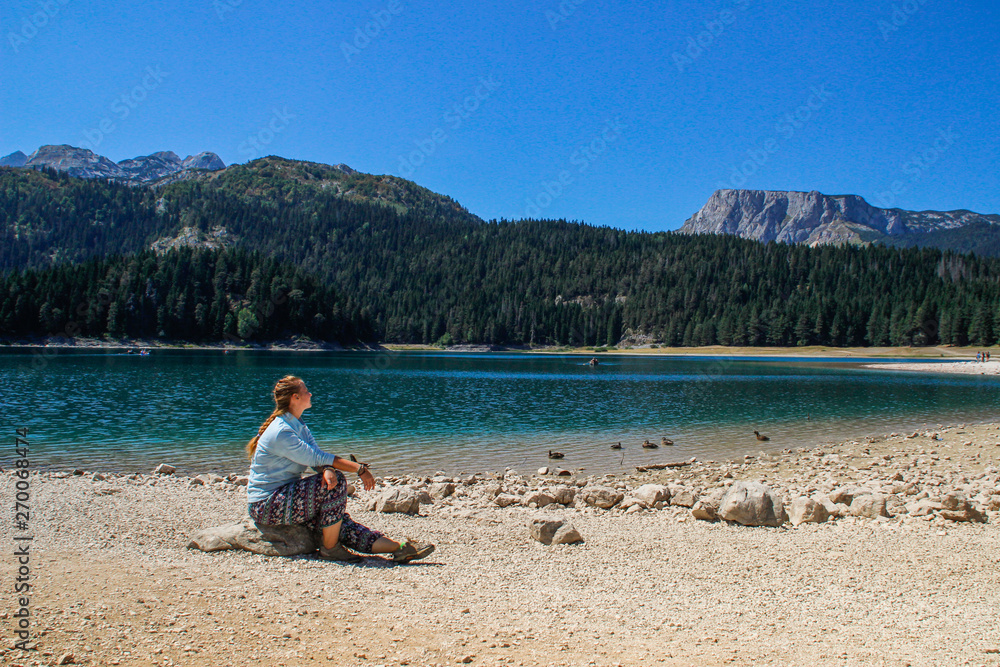 Paradise views of the national park Durmitor in Montenegro. Turquoise water of the lake, pine forest and mountains. Stunning background with nature girl tourist sitting on the beach Paradise views of the national park Durmitor in Montenegro. Turquoise water of the lake, pine forest and mountains. Stunning background with nature girl tourist sitting on the beach