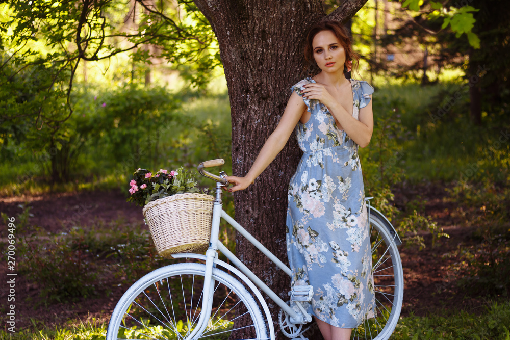 portrait of a beautiful girl in the forest, holding a bike with a basket of flowers, behind the rays of the sun, a blue flowered dress, summer walk portrait of a beautiful girl in the forest, holding a bike with a basket of flowers, behind the rays of the sun, a blue flowered dress, summer walk