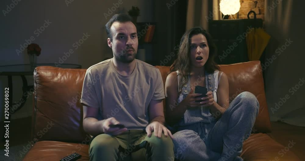 Young couple sits on couch and is distracted by TV program from texting on smartphones Young couple sits on couch and is distracted by TV program from texting on smartphones