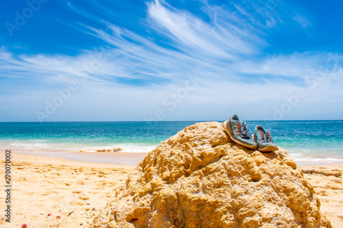 Pair of sport shoes lying on stone on beach against blue sky. Praia de Marinha in Algarve, Portugal 