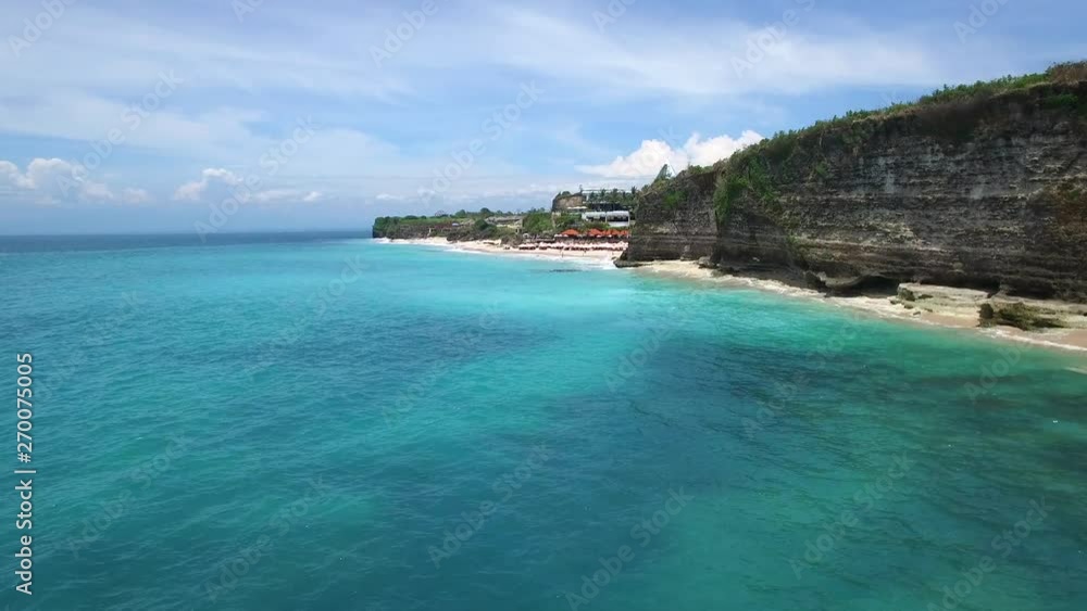 Long sandy beach on tropical island surrounded by cliffs with green vegetation on top and azure Indian Ocean with frothy waves on bright sunny day. Aerial footage of Dreamland beach, Bali, Indonesia. Long sandy beach on tropical island surrounded by cliffs with green vegetation on top and azure Indian Ocean with frothy waves on bright sunny day. Aerial footage of Dreamland beach, Bali, Indonesia.