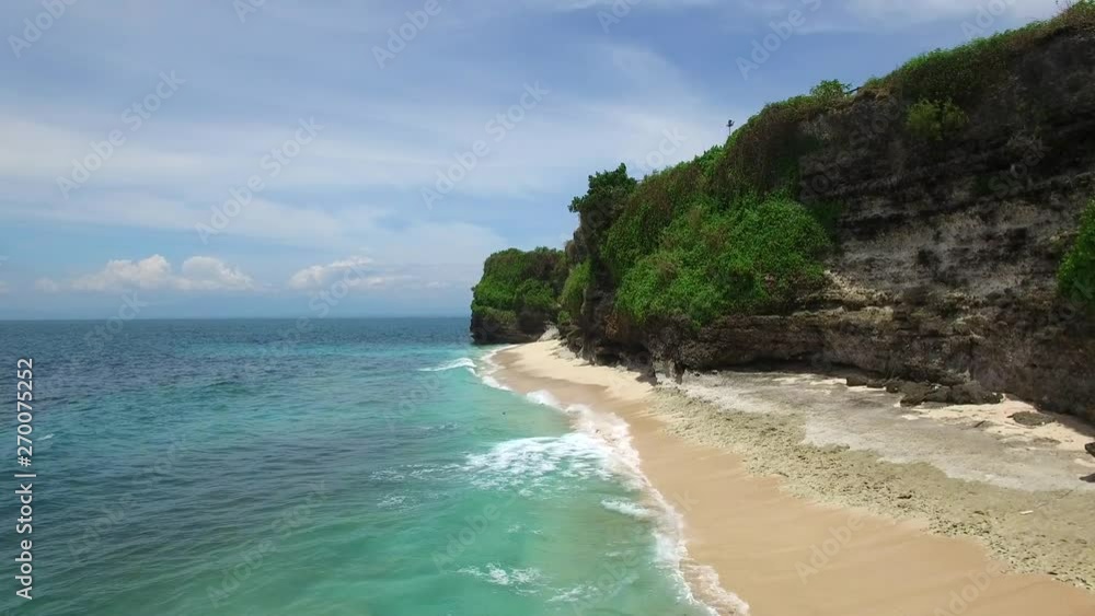Long sandy beach on tropical island surrounded by cliffs with green vegetation on top and azure Indian Ocean with frothy waves on bright sunny day. Aerial footage of Dreamland beach, Bali, Indonesia. Long sandy beach on tropical island surrounded by cliffs with green vegetation on top and azure Indian Ocean with frothy waves on bright sunny day. Aerial footage of Dreamland beach, Bali, Indonesia.