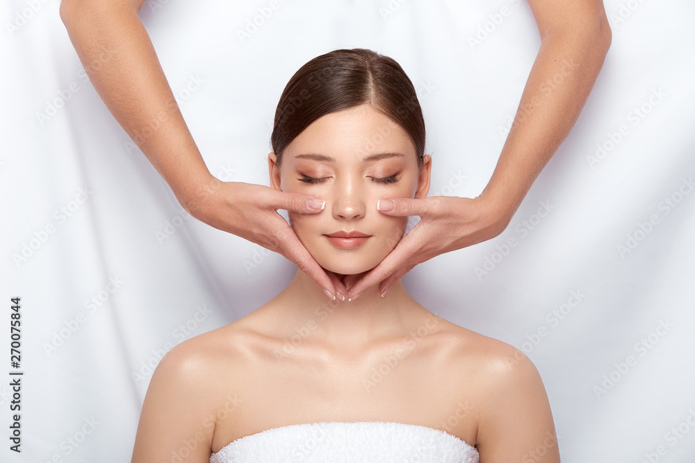 Naklejka premium attractive girl having facial massage with her eyes closed, natural skin care in spa salon, female arms doing face treatment