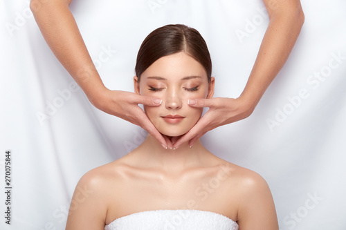 attractive girl having facial massage with her eyes closed, natural skin care in spa salon, female arms doing face treatment