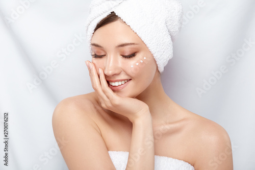cute girl in bath towel and cream on the cheek looking down with smile, young happy woman moisturizing her face