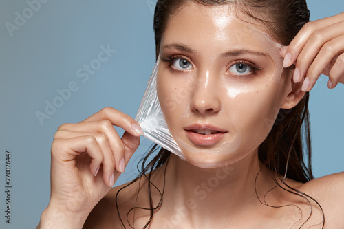 Obraz na płótnie close-up portrait of pretty girl taking off trasparent facial mask isolated on blue, cosmetology procedures, beauty and natural concept w salonie