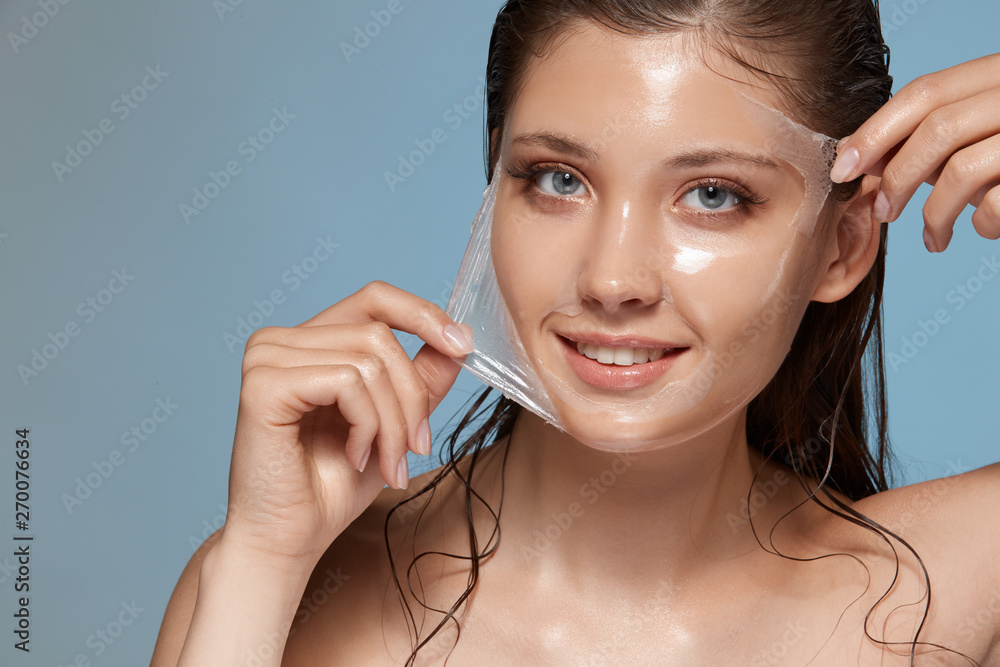 woman peels-off transparent facial mask and smiling to the camera on blue, happy anf fresh girl with perfect skin