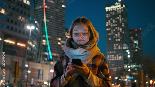 Caucasian Young Woman is Using Smartphone App Outdoors in the Downtown City in the Late Evening or Night with Beautiful Blurred Cityscape Lights of Skyscraper Windows