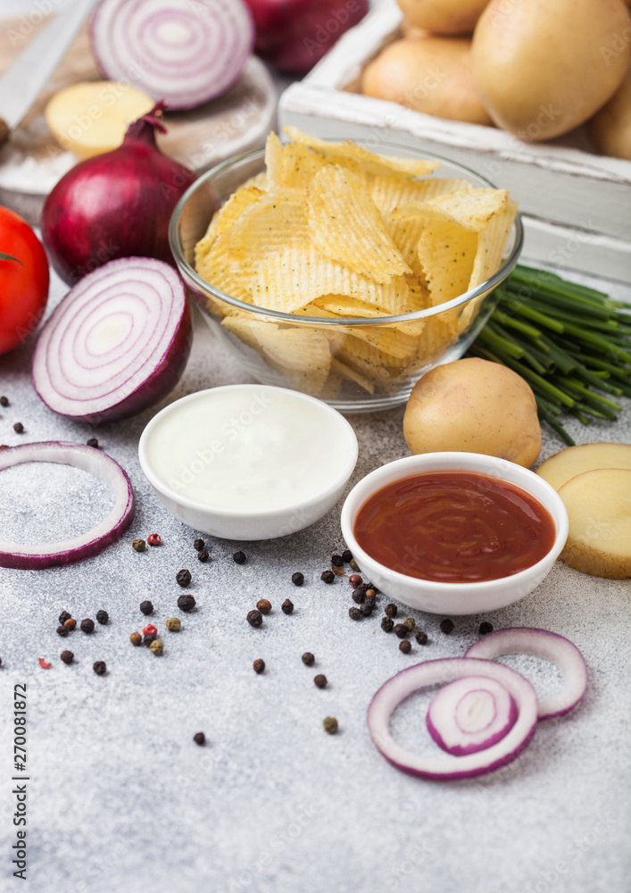 Fresh organic homemade potato crisps chips in glass bowl with sour cream and red onions on light kitchen table background. Box of fresh yellow potatoes, bowl of ketchup and green onion and tomatoes Fresh organic homemade potato crisps chips in glass bowl with sour cream and red onions on light kitchen table background. Box of fresh yellow potatoes, bowl of ketchup and green onion and tomatoes