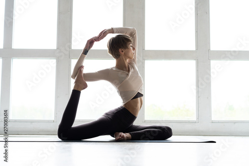 Young attractive smiling woman practicing yoga, sitting in One Legged King Pigeon exercise, Eka Pada Rajakapotasana pose, working out, wearing sportswear, brown pants, bra, indoor full length, home.