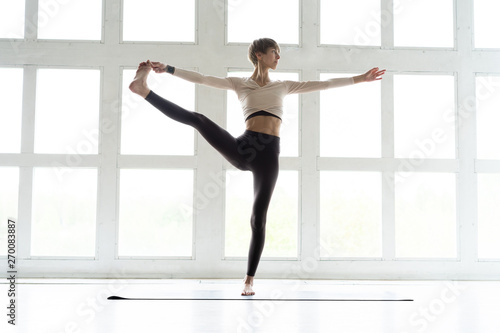 Portrait of attractive happy young woman working out indoors, doing yoga exercise, variation of Natarajasana, Lord of the Dance, King Dancer or Standing Mermaid Pose.