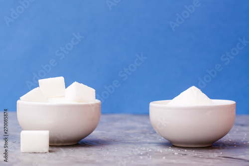 Sugar cubes and granulated sugar in white bowls on a table against a blue background. Concept photo