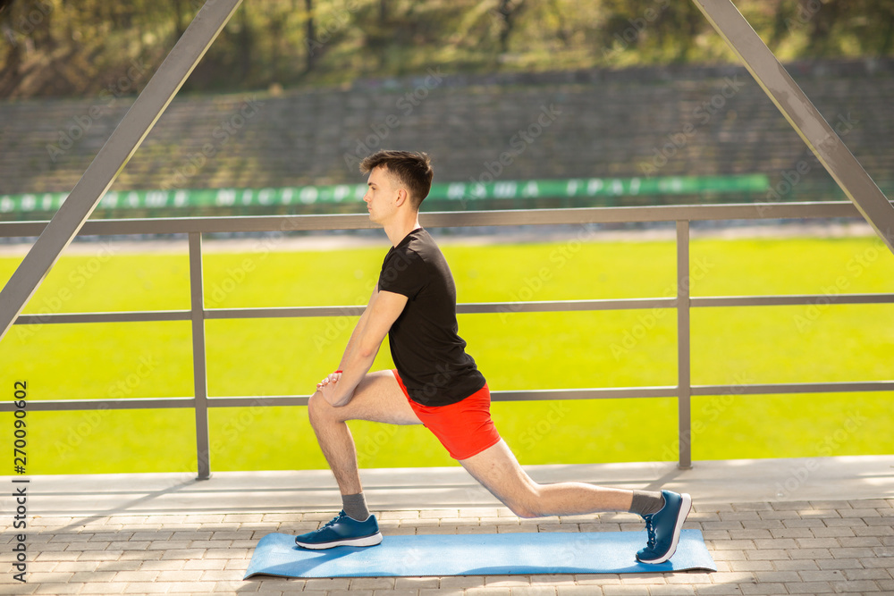 Young man training yoga outdoors. Sporty guy makes stretching exercise on a blue yoga mat, on the sports ground.