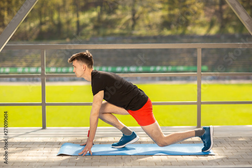 Young man training yoga outdoors. Sporty guy makes stretching exercise on a blue yoga mat, on the sports ground.