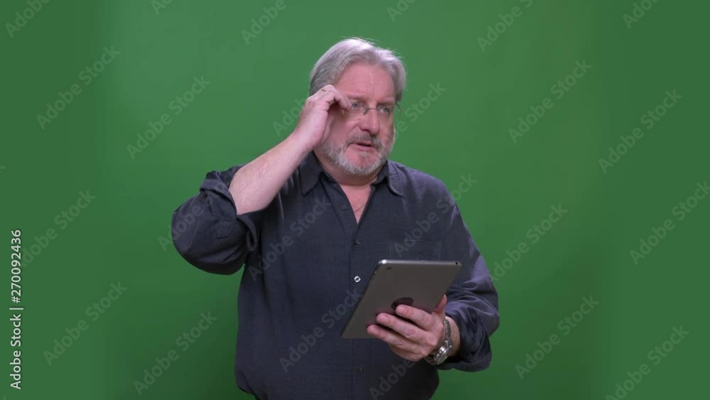 Senior gray haired businessman takes off glasses working with tablet isolated on green chromakey background. Senior gray haired businessman takes off glasses working with tablet isolated on green chromakey background.