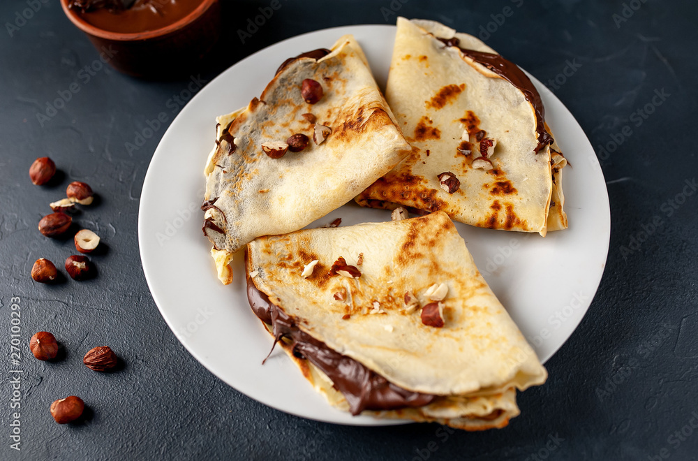 Pancakes with chocolate paste and hazelnuts, on a white plate on a background of concrete, slate Pancakes with chocolate paste and hazelnuts, on a white plate on a background of concrete, slate