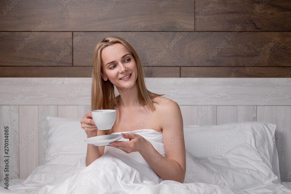 An attractive middle-aged blonde woman in a white bathrobe is lying on a bed in a bedroom. Drinking coffee from a white cup. Smiles and laughs. An attractive middle-aged blonde woman in a white bathrobe is lying on a bed in a bedroom. Drinking coffee from a white cup. Smiles and laughs.
