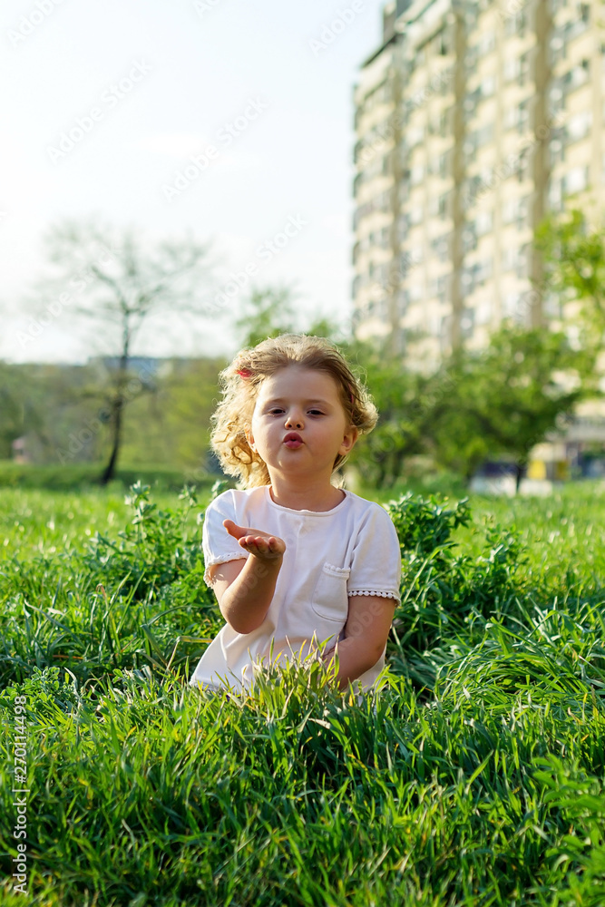 Fototapeta premium Cute little girl sending a kiss, sitting in the grass, on a sunny spring day